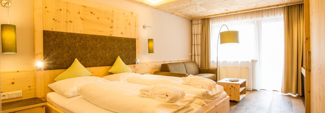 Ein gemütliches Hotelzimmer mit Holzdekor. Es enthält ein Doppelbett, einen kleinen Sitzbereich und große Fenster mit viel Licht. - Bäckelar Wirt - Sölden
