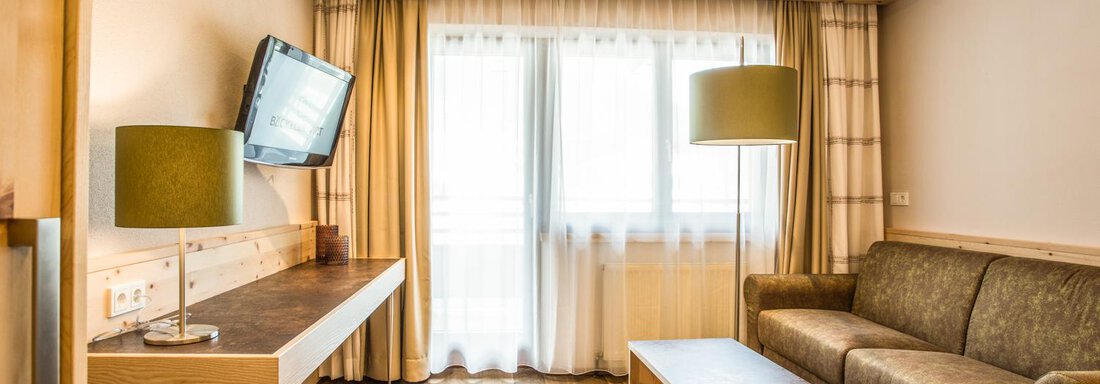 Ein modernes Wohnzimmer mit einem Sofa und einem Holztisch. Der Raum hat große Fenster mit Vorhängen und eine Wandmontage für einen Fernseher. - Bäckelar Wirt - Sölden