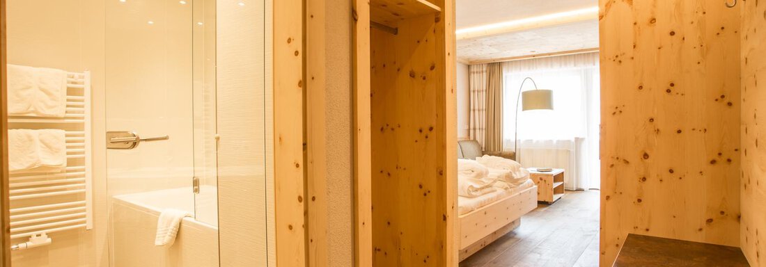 Ein modernes Hotelzimmer mit warmen Holztönen. Das Badezimmer ist durch eine Glaswand abgetrennt und verfügt über eine Badewanne. - Bäckelar Wirt - Sölden