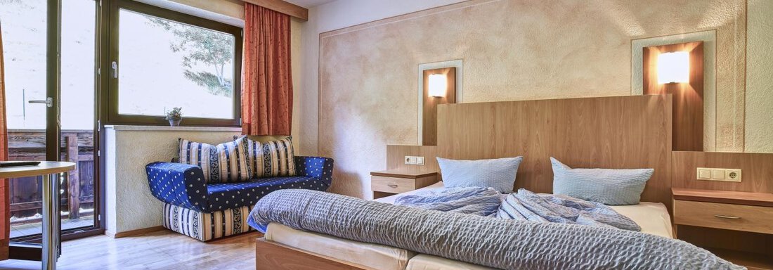 A cozy bedroom with a double bed and blue pillows. There is a small living area with a sofa and a balcony window. - Belledonne Ski in & out Appartements - Sölden