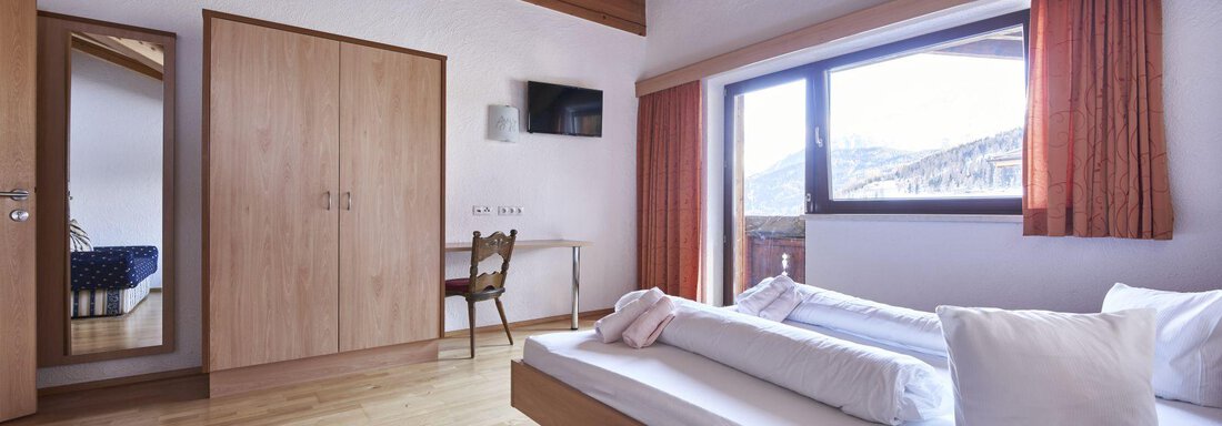 A bright room with wooden ceilings and large windows. There is a double bed, a table, and a mirror. - Belledonne Ski in & out Appartements - Sölden