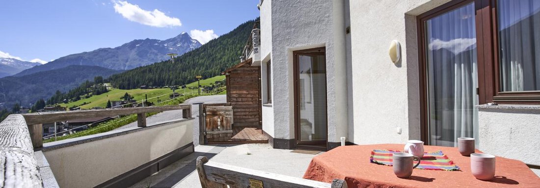A balcony with a view of the mountains and a cozy seating area. The sky is clear and the landscape is green. - Belledonne Ski in & out Appartements - Sölden