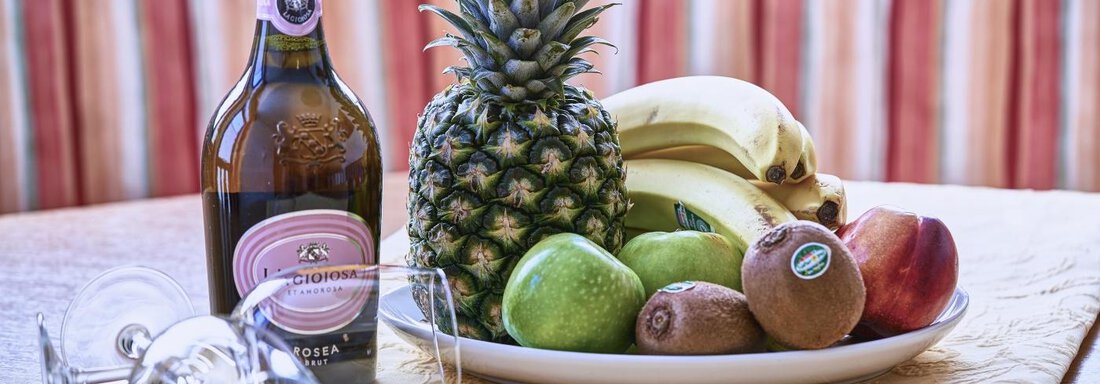 A bottle of sparkling wine stands next to a selection of fresh fruits, including pineapples, bananas, apples, and kiwis. The table is set with glasses and a decorative tablecloth. - Belledonne Ski in & out Appartements - Sölden