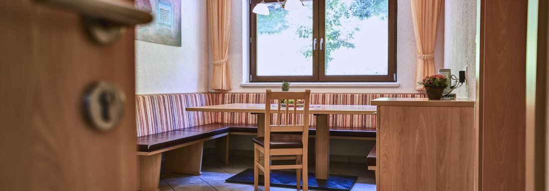 A cozy dining area with a wooden bench and table. Daylight streams through the window, and there are curtains in soft colors. - Belledonne Ski in & out Appartements - Sölden