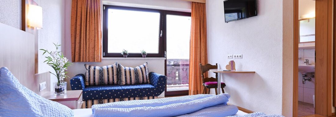 A bright hotel room with a double bed and blue bedspreads. There is a sofa, a television, and a window overlooking the outside. - Belledonne Ski in & out Appartements - Sölden