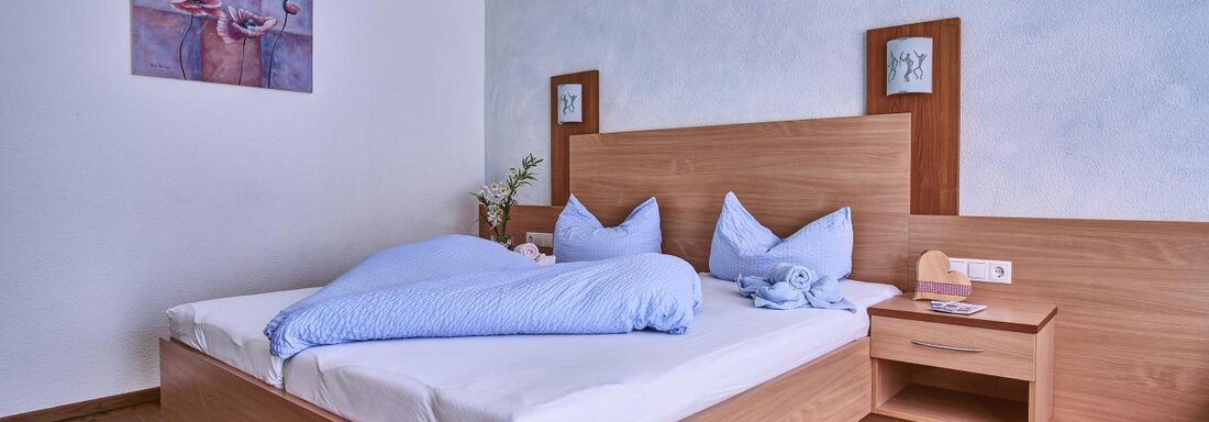 A bright bedroom with a double bed and blue pillows. A piece of artwork hangs on the wall, and a nightstand stands next to the bed. - Belledonne Ski in & out Appartements - Sölden