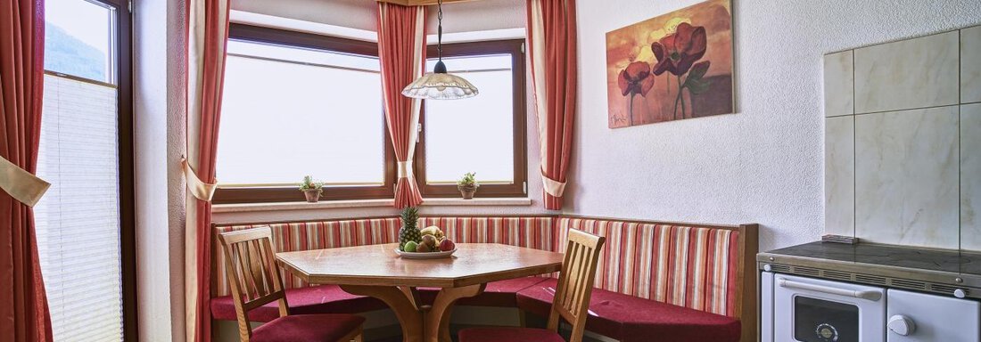 A cozy kitchen with a round table and wooden chairs. Large windows provide plenty of light, and red curtains create a warm atmosphere. - Belledonne Ski in & out Appartements - Sölden