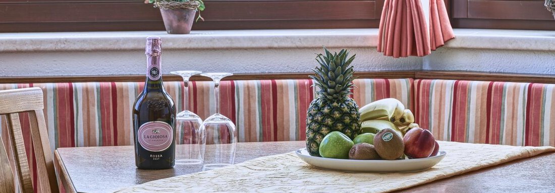 A cozy table with a bottle of wine, glasses, and fresh fruit, including a pineapple, apples, and a banana. In the background, a well-decorated windowsill with plants can be seen. - Belledonne Ski in & out Appartements - Sölden