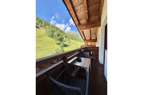 A cozy balcony with two chairs and a small table. In the background, green mountains and a blue sky can be seen. - Belledonne Ski in & out Appartements - Sölden
