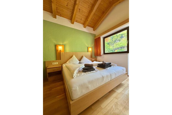 A modern bedroom with wood details and a large window. The bed is neatly made and some towels are lying on the bedspread. - Belledonne Ski in & out Appartements - Sölden