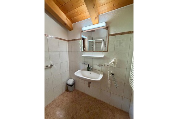 A modern bathroom with white tiles and a wooden ceiling. There is a sink, a mirror, and a shower. - Belledonne Ski in & out Appartements - Sölden