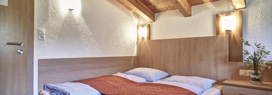 A cozy bedroom with wooden ceilings and a double bed. The walls are painted light and there is a small flower on the nightstand. - Belledonne Ski in & out Appartements - Sölden