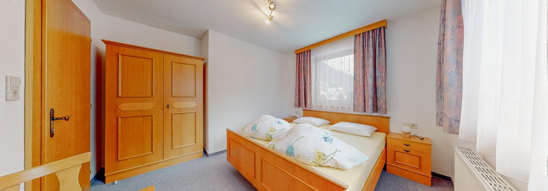 A simple bedroom with a double bed and wood-colored furniture. The walls are painted light and large windows provide daylight. - Belmonte Sölden - Sölden