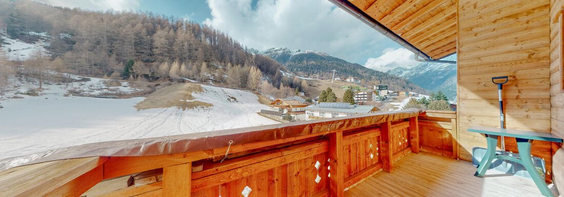 A wooden-clad balcony with a view of snow-covered mountains. The clouds drift past in the blue sky and some buildings are visible in the valley. - Belmonte Sölden - Sölden