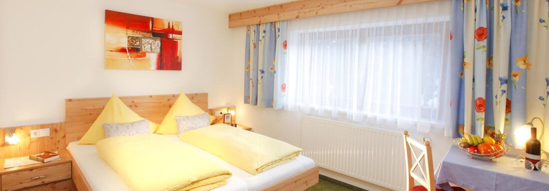 A cozy bedroom with a double bed and yellow pillows. The room is bright and friendly, with a table and chairs as well as colorful curtains. - Brandlehof - Sölden