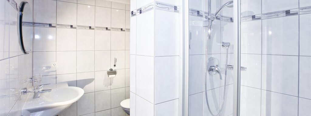 A modern bathroom with white tiles and a shower. There is a sink and a toilet, all looking clean and well-equipped. - Brandlehof - Sölden