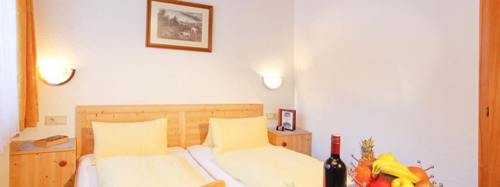 A cozy room with two single beds and wooden decor. On the table, there is a bottle of wine and a bowl of fresh fruits. - Brandlehof - Sölden