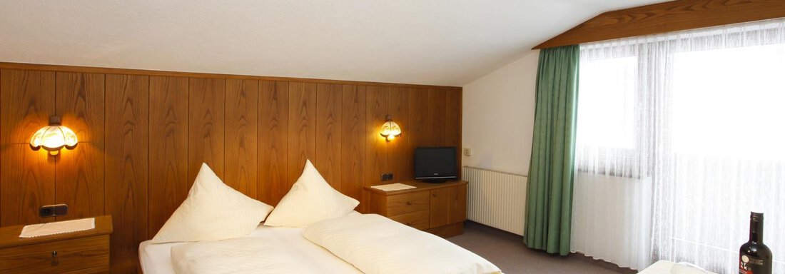 A cozy bedroom with a double bed and wooden paneling. There is a table with a bottle of wine and a television in the room. - Brandlehof - Sölden