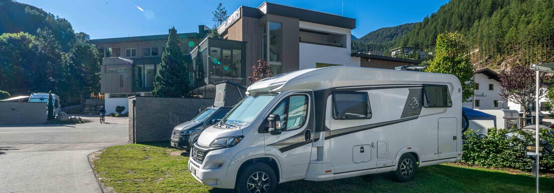 Stellplatz Komfort Sommer A motorhome is standing on a meadow in front of a modern building. In the background, green hills and a clear, blue sky are visible. - Camping Sölden - Sölden