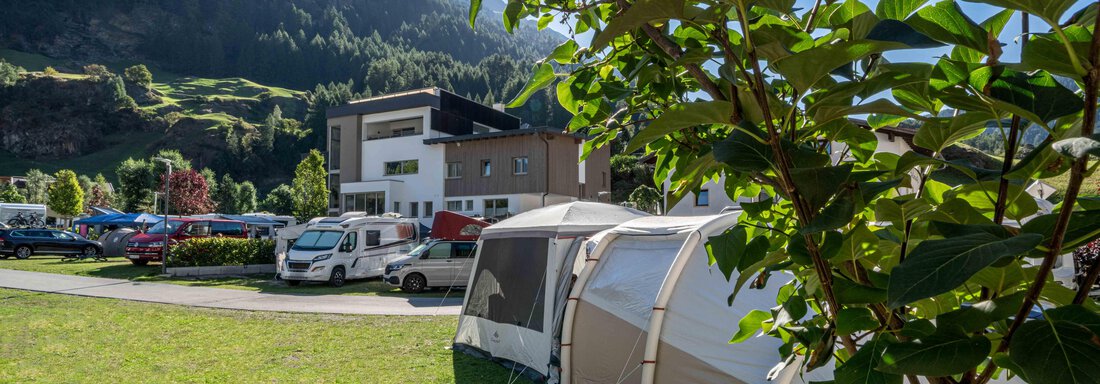 Stellplatz Premium A campsite with tents and campers in the foreground. In the background, green mountains and a modern building can be seen. - Camping Sölden - Sölden