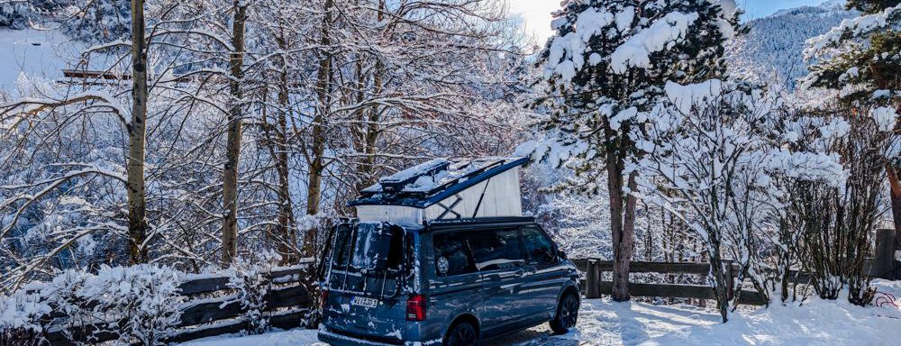 Ein blauer Campervan steht in einer schneebedeckten Landschaft. Hohe Bäume und ein klarer Himmel ergänzen die winterliche Szene. - Camping Sölden - Sölden