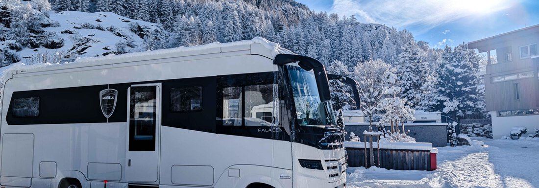 CS_Platz-Winter-komfort Ein Wohnmobil steht in verschneiter Umgebung unter blauem Himmel. Im Hintergrund sind schneebedeckte Bäume und Hügel zu sehen. - Camping Sölden - Sölden