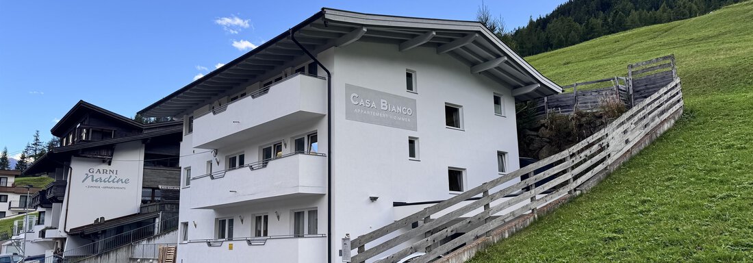 IMG_6383 A modern building with several floors is situated on the hillside. In the background, green meadows and mountains can be seen under a clear blue sky. - CASA bianco DUE - Sölden