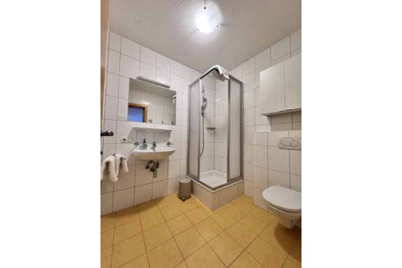 A modern bathroom with a shower cabin, sink, and toilets. The walls are tiled in a light color, and the floor is in a warm tone. - CASA bianco DUE - Sölden
