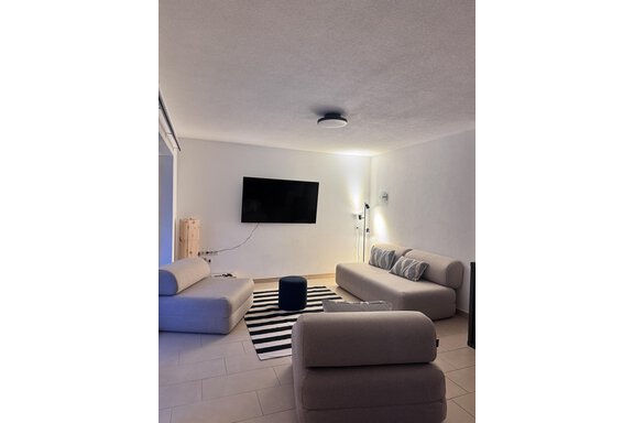 A modern living room with beige sofas and a blue pouf. There is a large television on the wall, and the floor has a black-and-white striped carpet. - CASA bianco DUE - Sölden