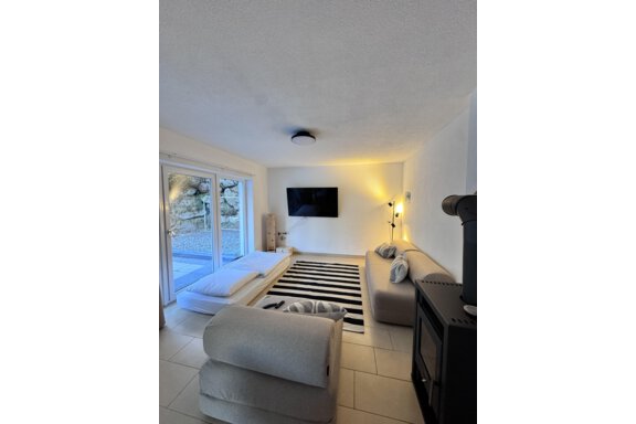 A modern living room with a cozy couch and a black carpet. Large windows let in plenty of light, and a television is mounted on the wall. - CASA bianco DUE - Sölden