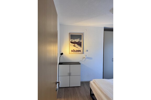 A cozy bedroom with a modern nightstand and a large wardrobe. A framed poster of Sölden is hanging on the wall. - CASA bianco DUE - Sölden