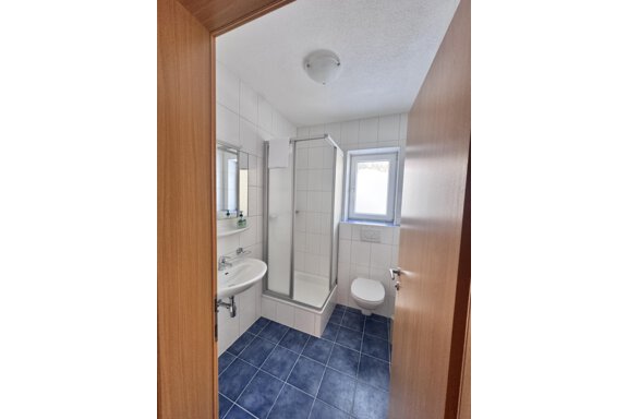 A modern bathroom with a shower, a sink, and a toilet. The walls are light tiled and the floor is blue. - CASA bianco DUE - Sölden