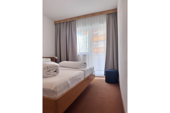 A simple bedroom with a bed, two towels, and a blue stool. Light curtains hang in front of the window, allowing daylight to come in. - CASA bianco DUE - Sölden
