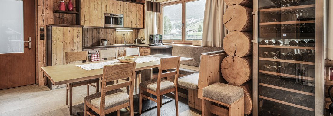 A cozy kitchen with wooden furniture and a large table. A wine cabinet stands against the wall and large windows provide plenty of natural light. - Chalet Resort Sölden - Sölden