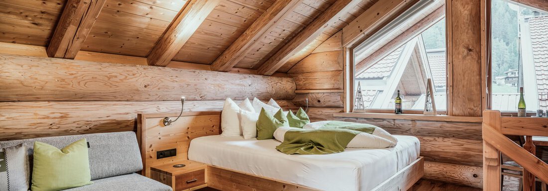 A cozy bedroom with wooden walls and sloping ceilings. There is a large bed, a seating area, and windows with a view of nature. - Chalet Resort Sölden - Sölden