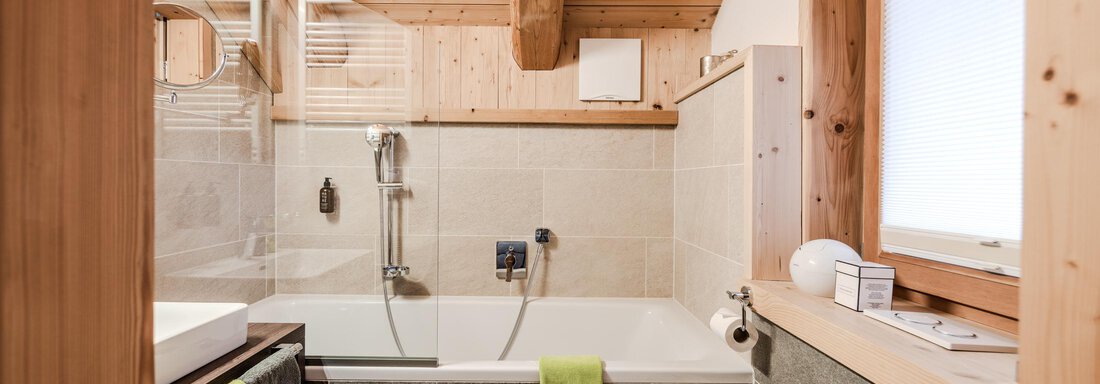 A modern bathroom with wooden elements and a window. The bathtub is equipped with gray tiles and towels in various colors. - Chalet Resort Sölden - Sölden