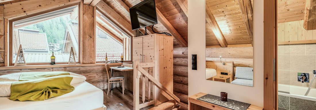 A cozy room with wood paneling and sloping ceilings. It is equipped with a bed, a television, and a bathroom. - Chalet Resort Sölden - Sölden