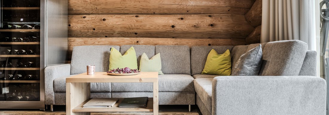 A modern living room with a gray couch and colorful cushions. A wooden table is in front of the couch, and a wine cabinet is visible in the background. - Chalet Resort Sölden - Sölden