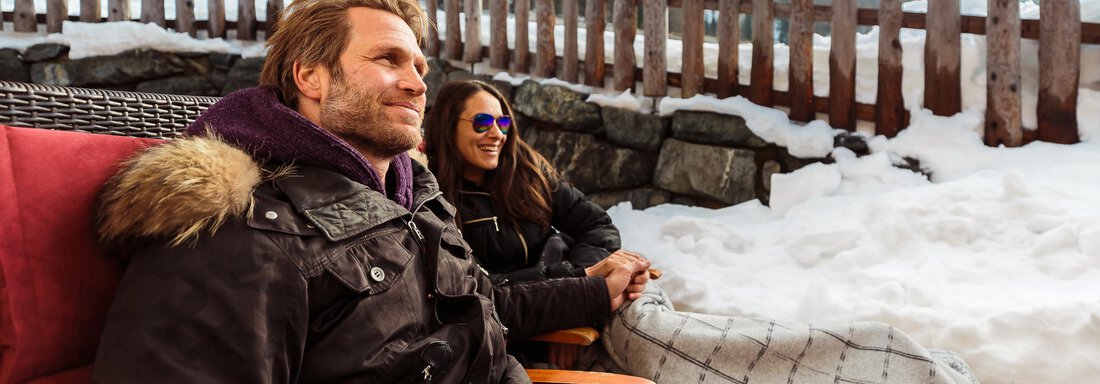 A couple sits in a cozy setting in the snow and enjoys their time together. They have a blanket and warm clothes while holding hot drinks. - Chalet Resort Sölden - Sölden