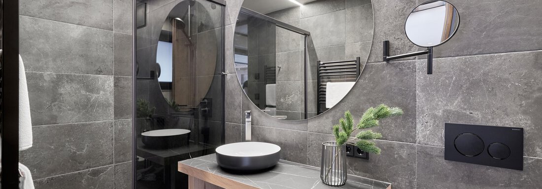 A modern bathroom with gray tiles and a glass shower. A round mirror and a stylish vanity top complement the elegant design. - Chalet White Pearl by MYALPS - Sölden