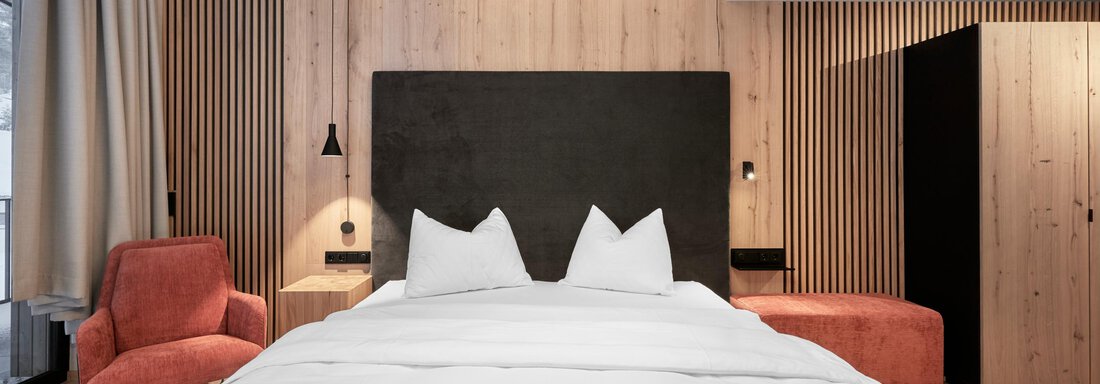 A modern bedroom with a large bed and a wooden panel. The stylish decor includes a red armchair and elegant lighting. - Chalet White Pearl by MYALPS - Sölden