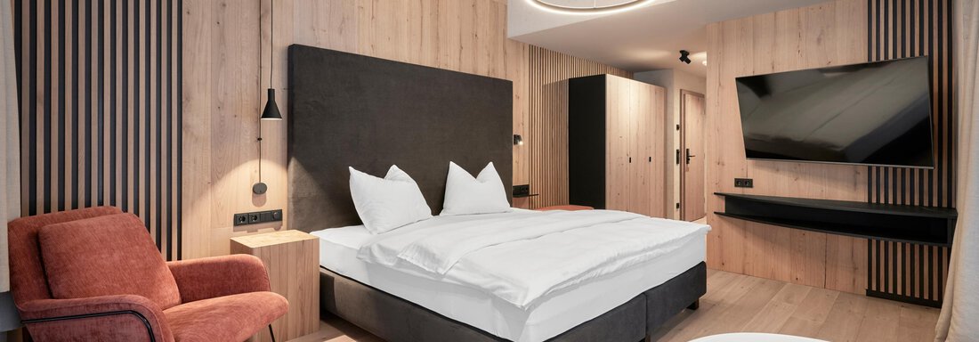 A modern bedroom with wooden paneling and a comfortable bed. In the room, there is a television set and a red armchair. - Chalet White Pearl by MYALPS - Sölden