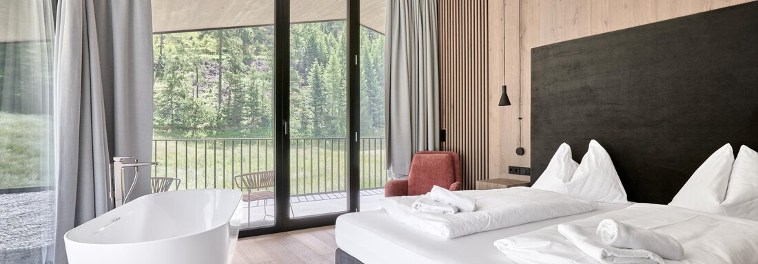 A modern room with a large window and a view of greenery. The furnishings include a double bed, a comfortable armchair, and a freestanding bathtub. - Chalet White Pearl by MYALPS - Sölden
