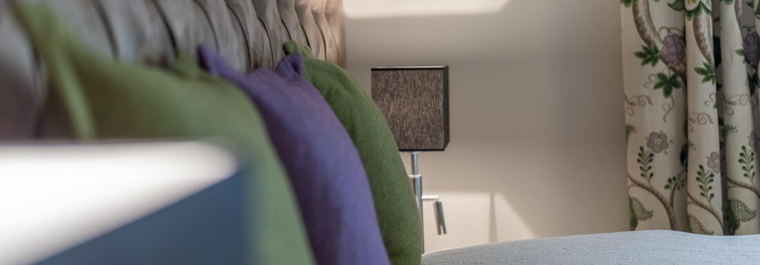 Ein gemütliches Schlafzimmer mit einem gepolsterten Kopfteil und bunten Kissen. Im Hintergrund sind Vorhänge mit floralem Muster zu sehen. - Das Central - Alpine . Luxury . Life - Sölden