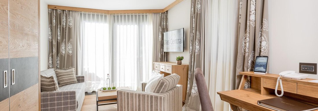 Ein modernes Wohnzimmer mit Holzfußboden und großen Fenstern. Die Einrichtung besteht aus stilvollen Möbeln und einem Schreibtisch. - Das Central - Alpine . Luxury . Life - Sölden