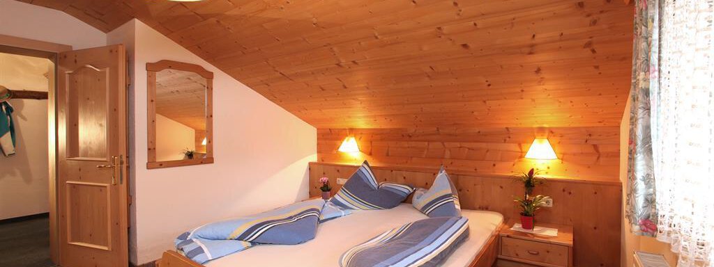A cozy wooden room with a double bed, blue bedspreads, and soft lighting. A large window with curtains creates an inviting atmosphere. - Erbhof - Sölden