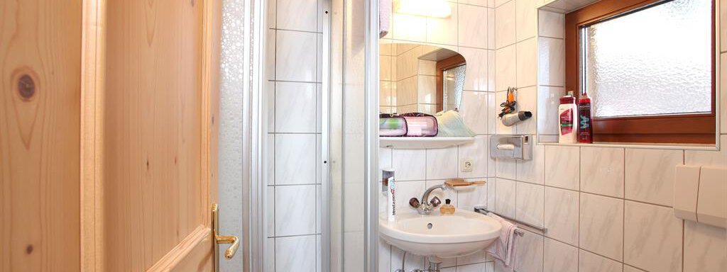 A small bathroom with a shower, sink, and toilet. The walls are tiled with white tiles and there is a window that lets in natural light. - Erbhof - Sölden