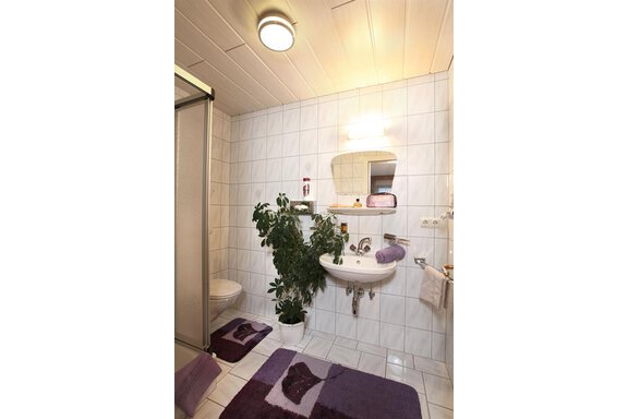 A modern bathroom with white tiles and a shower. There is a sink, plants, and a purple bath mat. - Erbhof - Sölden
