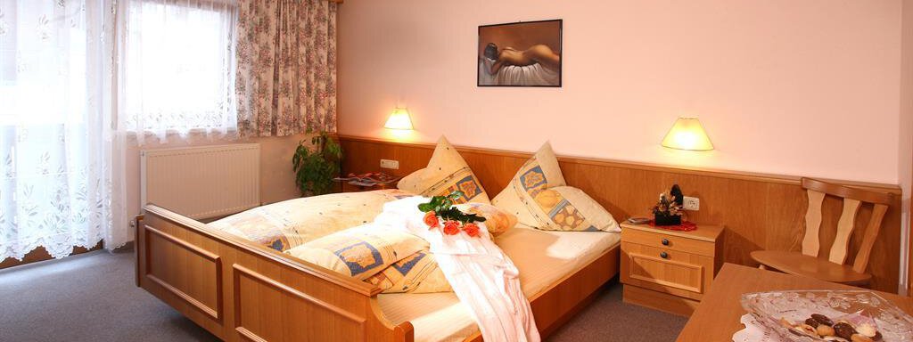 A cozy bedroom with a double bed, wooden furniture, and soft lighting. On the table, there is a bowl of snacks, and daylight filters in through the windows. - Erbhof - Sölden