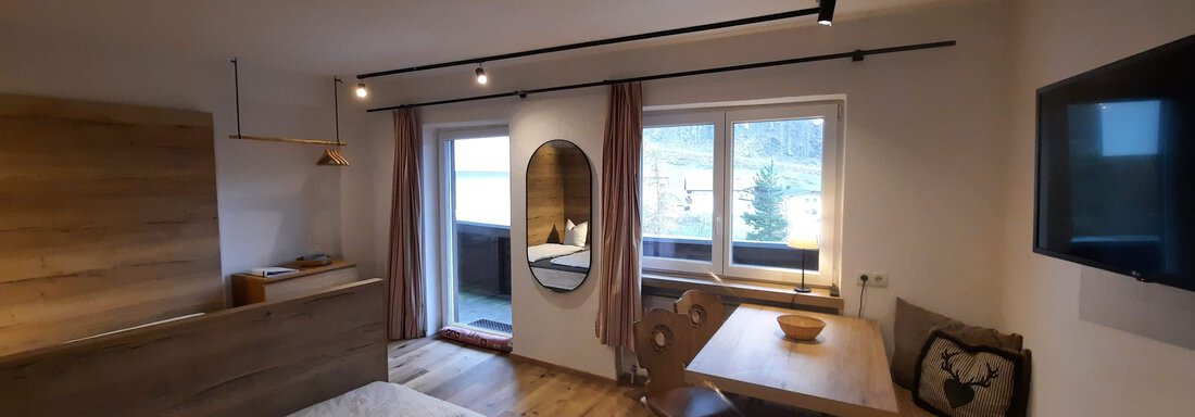 A modern room with wooden floors, a single bed, and a dining area. Large windows provide plenty of light and a view outside. - Ferienhaus Anna - Sölden